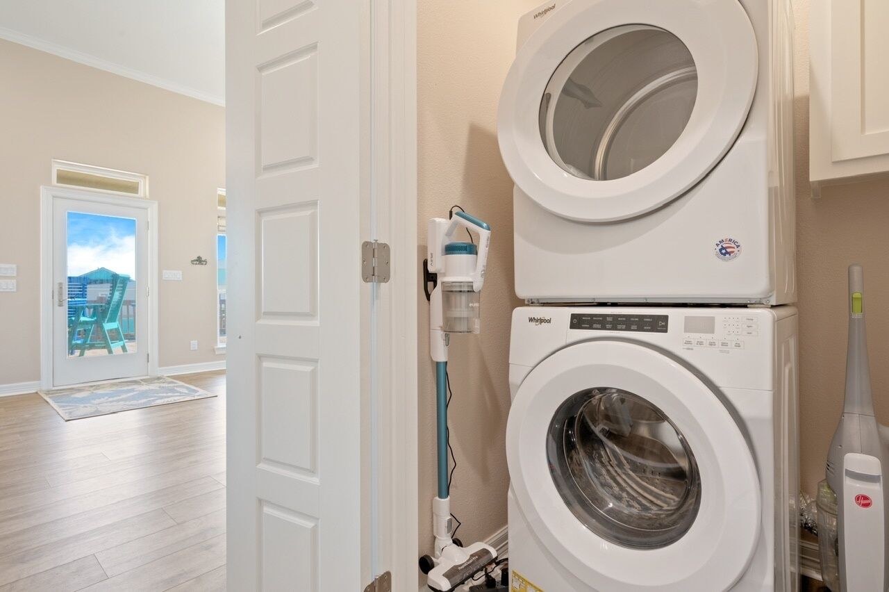 Une buanderie équipée d’une machine à laver et d’un sèche-linge superposés, d’un aspirateur et d’une porte donnant sur un balcon avec vue sur les immeubles.