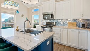 A modern kitchen with white cabinets, a central island with a sink, a stove, and a microwave. There are decorative starfish on the wall and a view of the outside through the window. - Nauti By Nature 3 Bedroom Home (Port Bolivar)