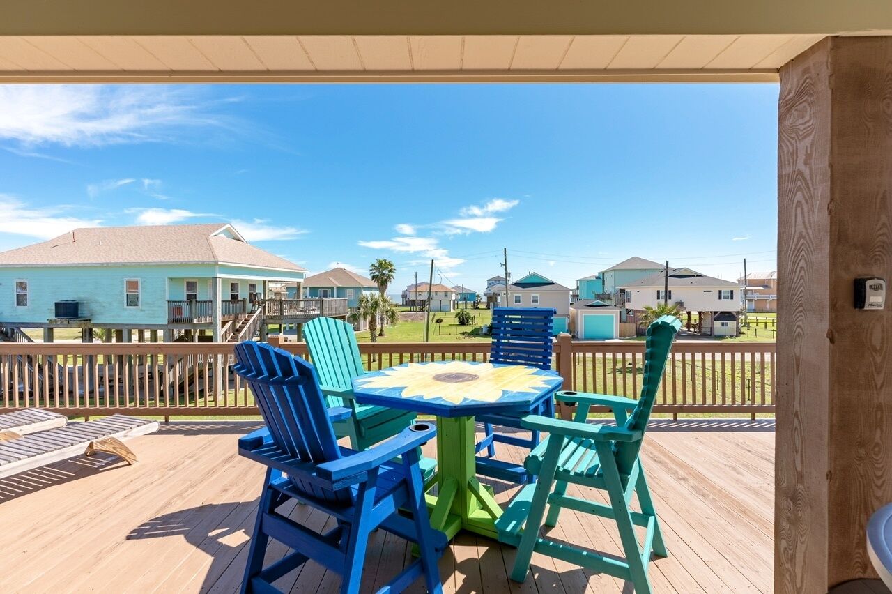 A wooden deck with a round table and chairs, a view of houses in the background, and a clear sky.