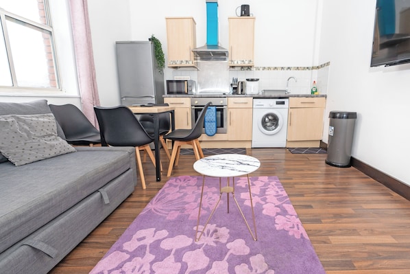 Un espacio habitable compacto con sofá, sillas, mesa de comedor, zona de cocina con electrodomésticos y televisor montado en la pared.