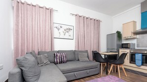Ein modernes Wohnzimmer mit einem grauen Ecksofa, zwei schwarzen Sesseln, einem violetten Teppich und einer Küchenzeile mit Mikrowelle, Backofen und Schränken.