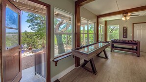 A pool table in a room with large windows, a foosball table, and a ceiling fan.
