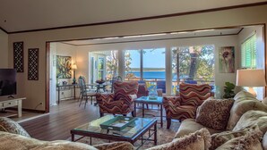 A spacious living room with a large sofa, armchairs, and a coffee table. There is a television on a stand, a dining table with chairs, and a view of the ocean through large windows.