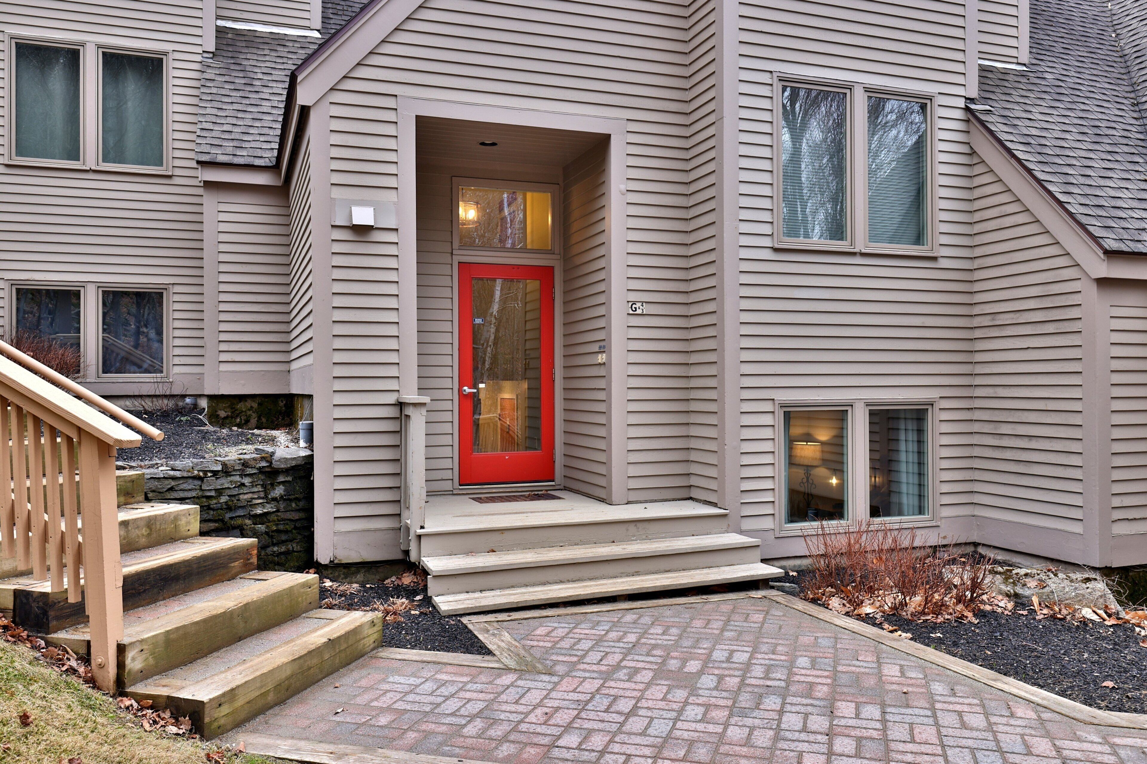 A two-story house with a red front door, a stone wall, and a brick pathway leading to the entrance.
