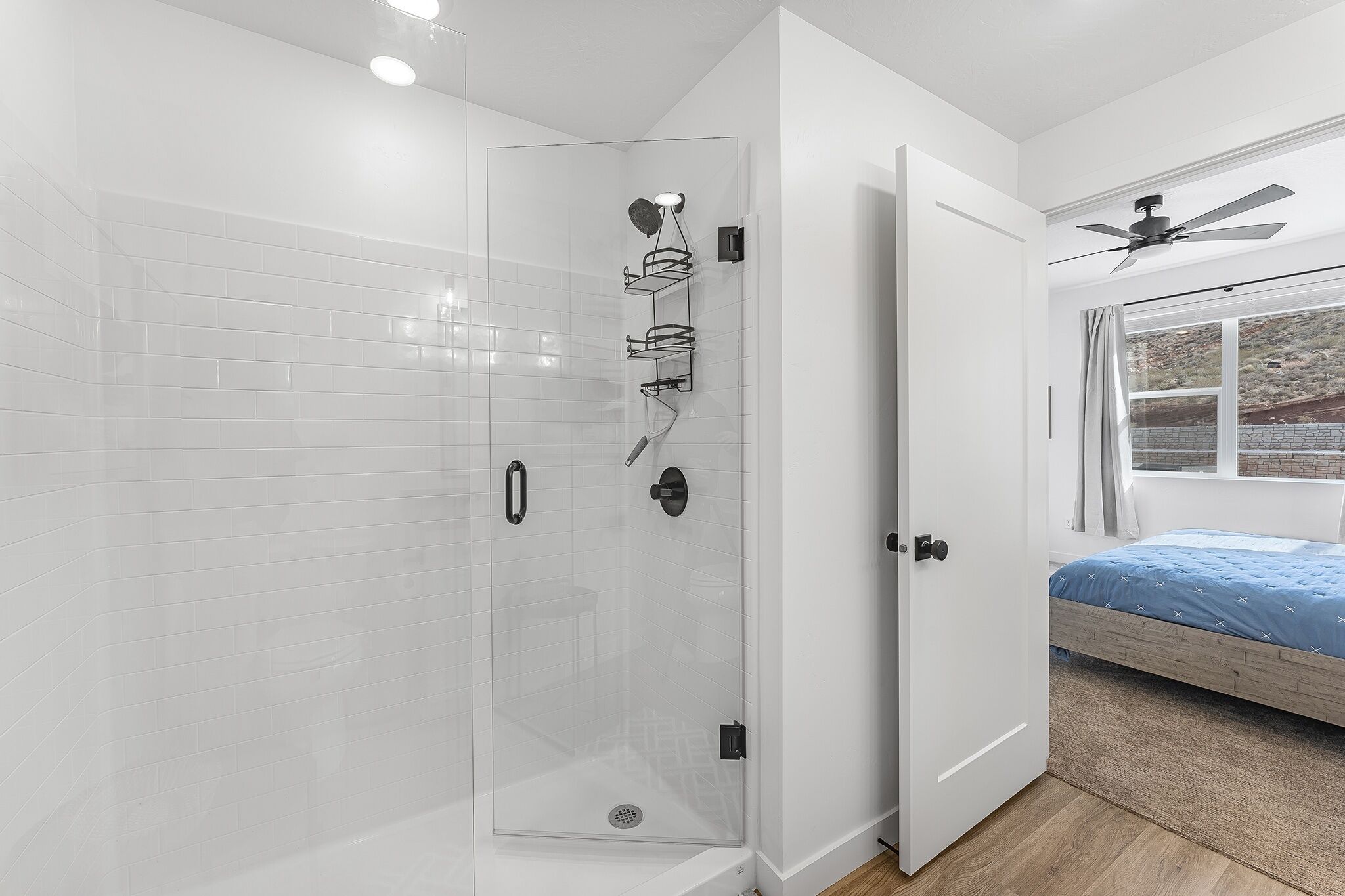 A modern bathroom with a glass-enclosed shower, a wooden bed with a blue bedspread, and a ceiling fan.