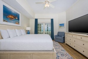 A hotel room with a large bed, a television, a desk with drawers, a chair, a ceiling fan, and a painting of a sea turtle.