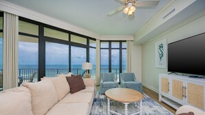 Amplia sala de estar con un sofá grande, una mesa de centro y dos sillones. Un televisor está montado en un mueble blanco y se ve un ventilador de techo. Amplias ventanas ofrecen vistas al océano.