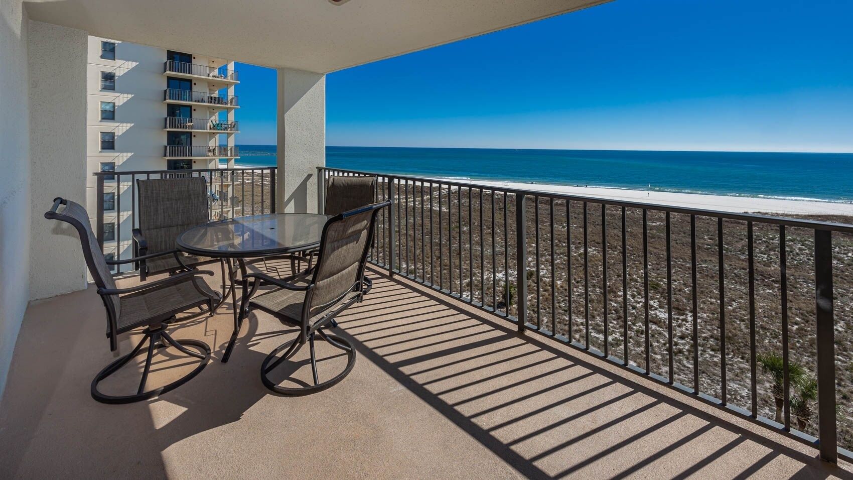Ein Balkon mit Glastisch und Stühlen, der Blick auf einen Strand und ein Gebäude mit mehreren Balkonen bietet.