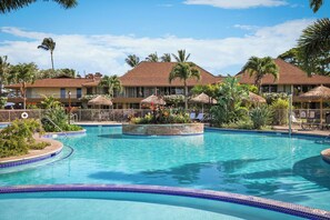 Ein Pool in einem Resort mit tropischer Umgebung, darunter Palmen und Reetdachschirme.