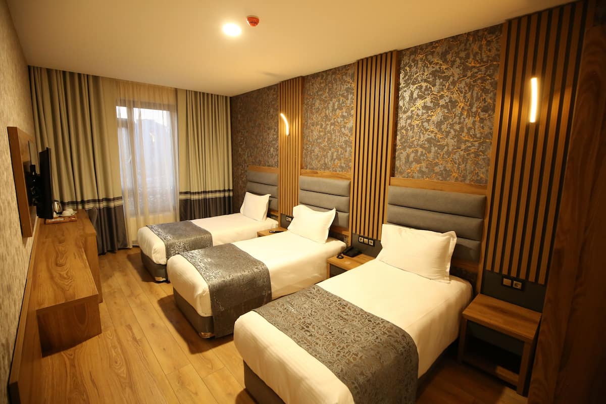 Quarto de hotel com três camas de solteiro, uma escrivaninha de madeira, televisão e uma janela grande com cortinas.