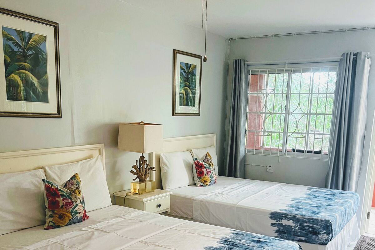 A hotel room with two beds, a ceiling fan, a window with curtains, and framed pictures on the wall.