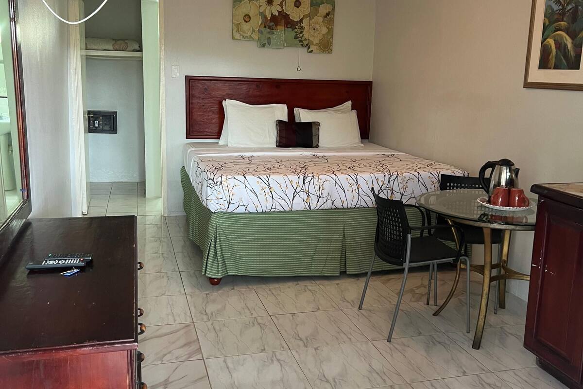 A hotel room with a bed, a ceiling fan, a TV mounted on the wall, a table with chairs, and a small kitchenette.