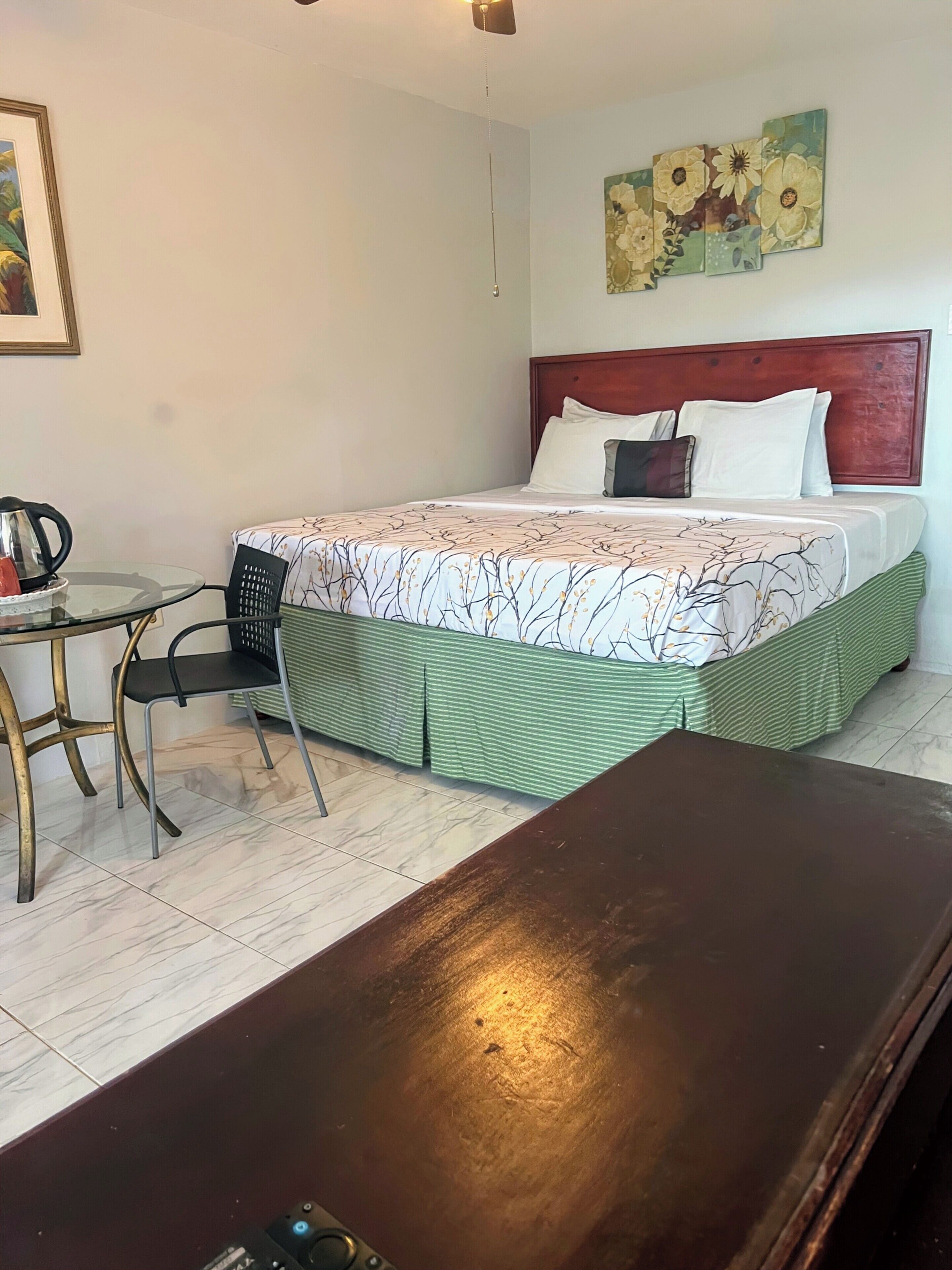 Habitación de hotel con cama, una mesita con una silla, un cuadro en la pared y un ventilador de techo.