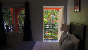 A hotel room with a bed, a window with a view of greenery, and a door leading outside. - Paradise On The Rocks Hotel (Negril)