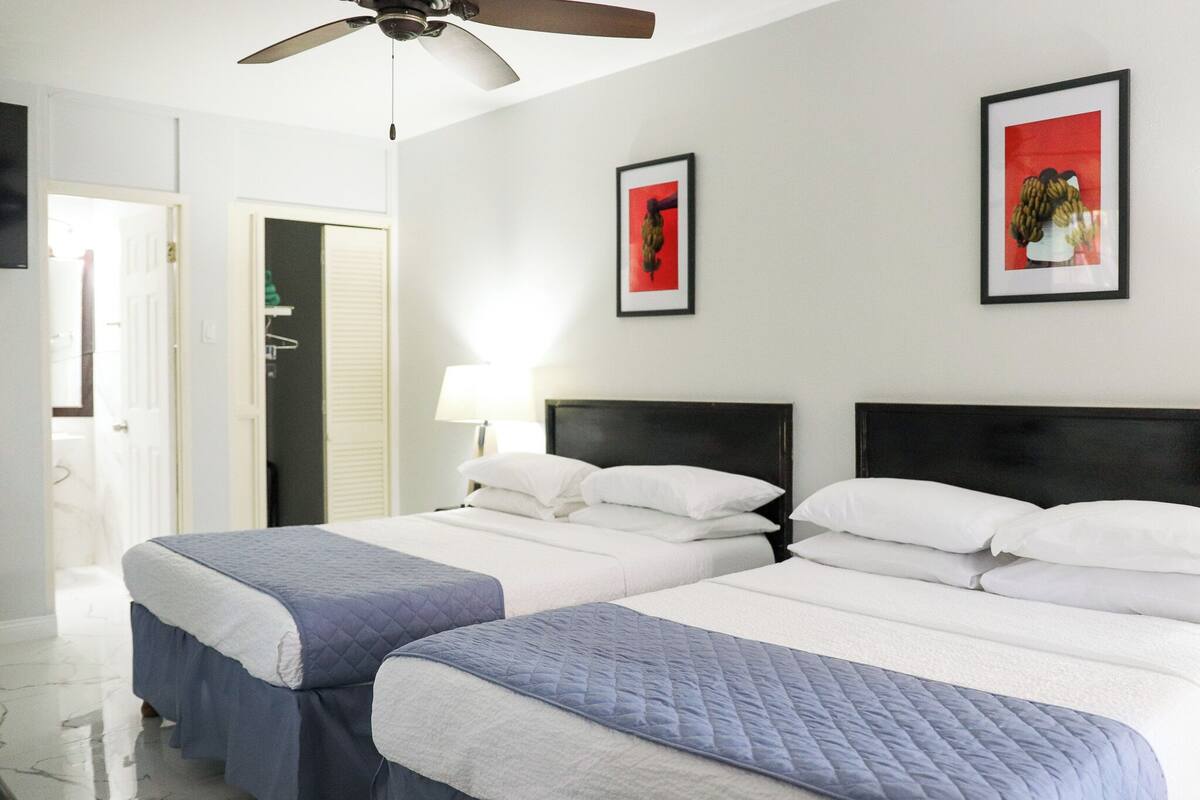 A hotel room with two beds, a ceiling fan, a door, and two framed pictures on the wall.