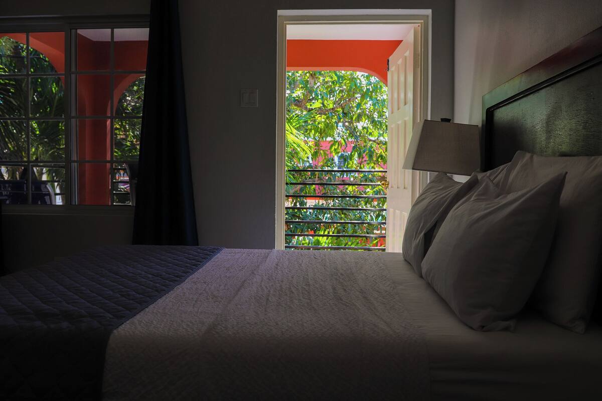 A hotel room with a bed, a nightstand, a lamp, and a view of a balcony with greenery.