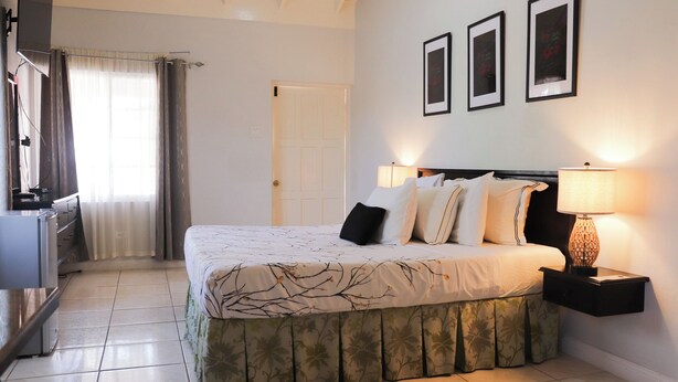 A neatly arranged bedroom with a bed, two bedside lamps, a nightstand, a wardrobe, and a door. - Paradise On The Rocks Hotel (Negril)