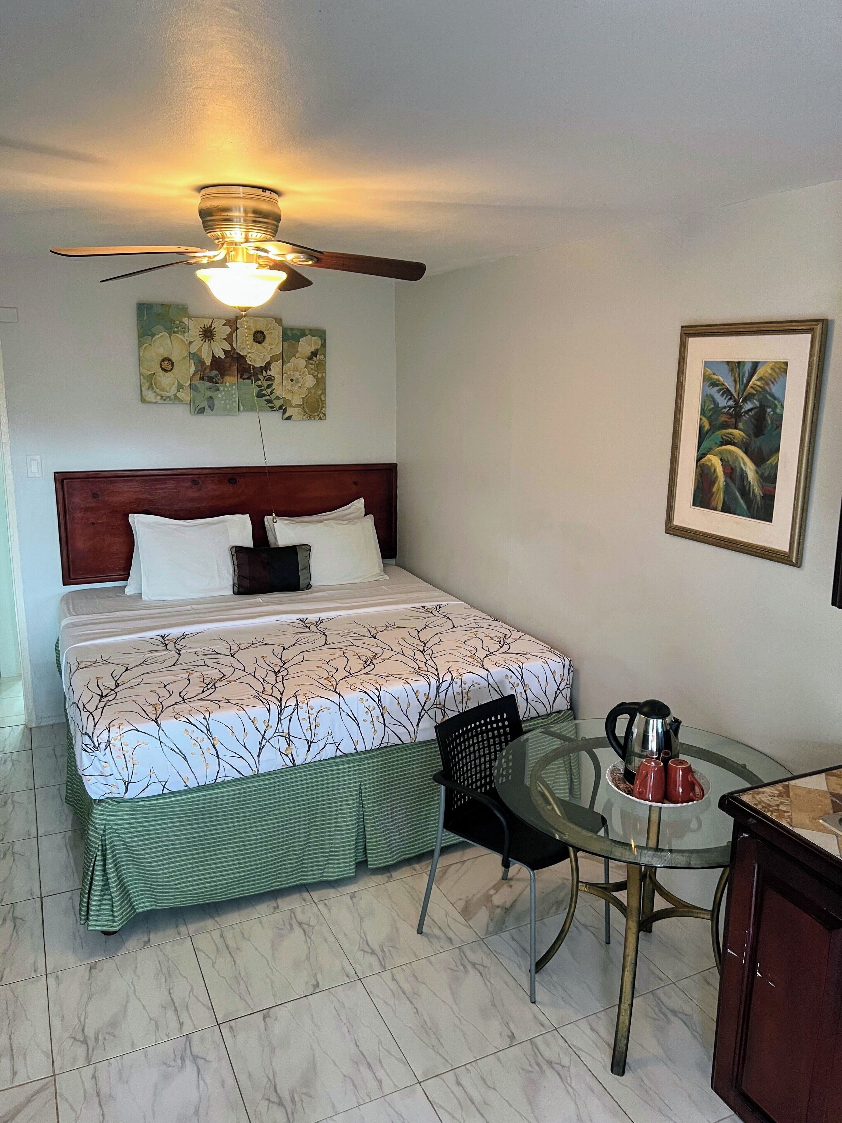 A neatly arranged bedroom with a bed, a ceiling fan, a wall-mounted light, a framed floral painting, a small table with a kettle and cups, and a chair.