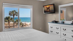 Quarto com uma janela grande que oferece vista para a praia, uma televisão de tela plana presa acima de uma cômoda branca e um ventilador de teto.