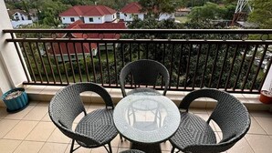 Un balcón con sillas de mimbre y una mesa de vidrio, con vista a una zona residencial con casas y una torre de líneas eléctricas.