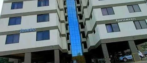 A modern apartment building with a central glass elevator shaft, multiple floors, and a parking garage at the base.