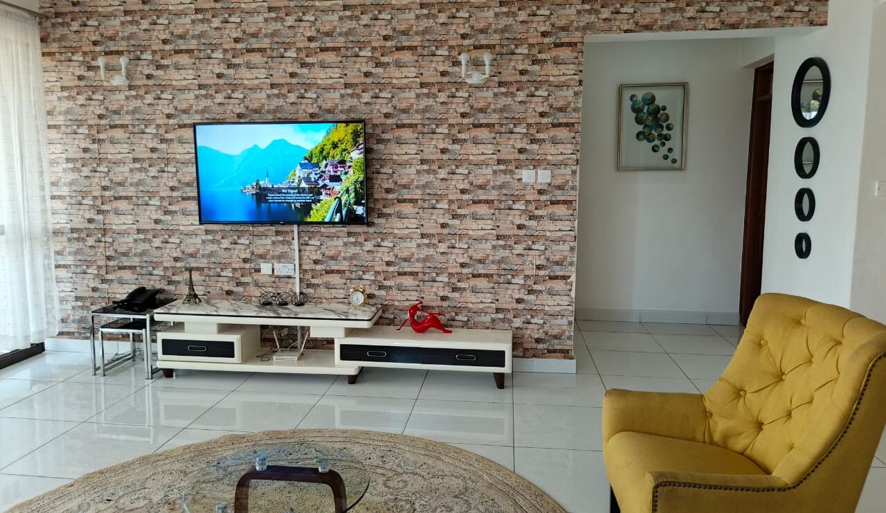 A living room with a brick wall, a flat-screen TV mounted above a wooden entertainment unit, a yellow armchair, and a patterned rug.