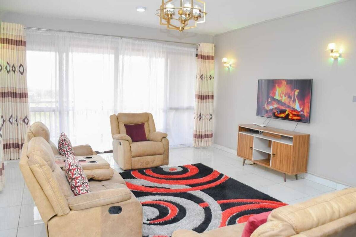 A living room with a beige sofa set, a red and black patterned rug, a wooden TV stand, and a painting of a fire on the wall.
