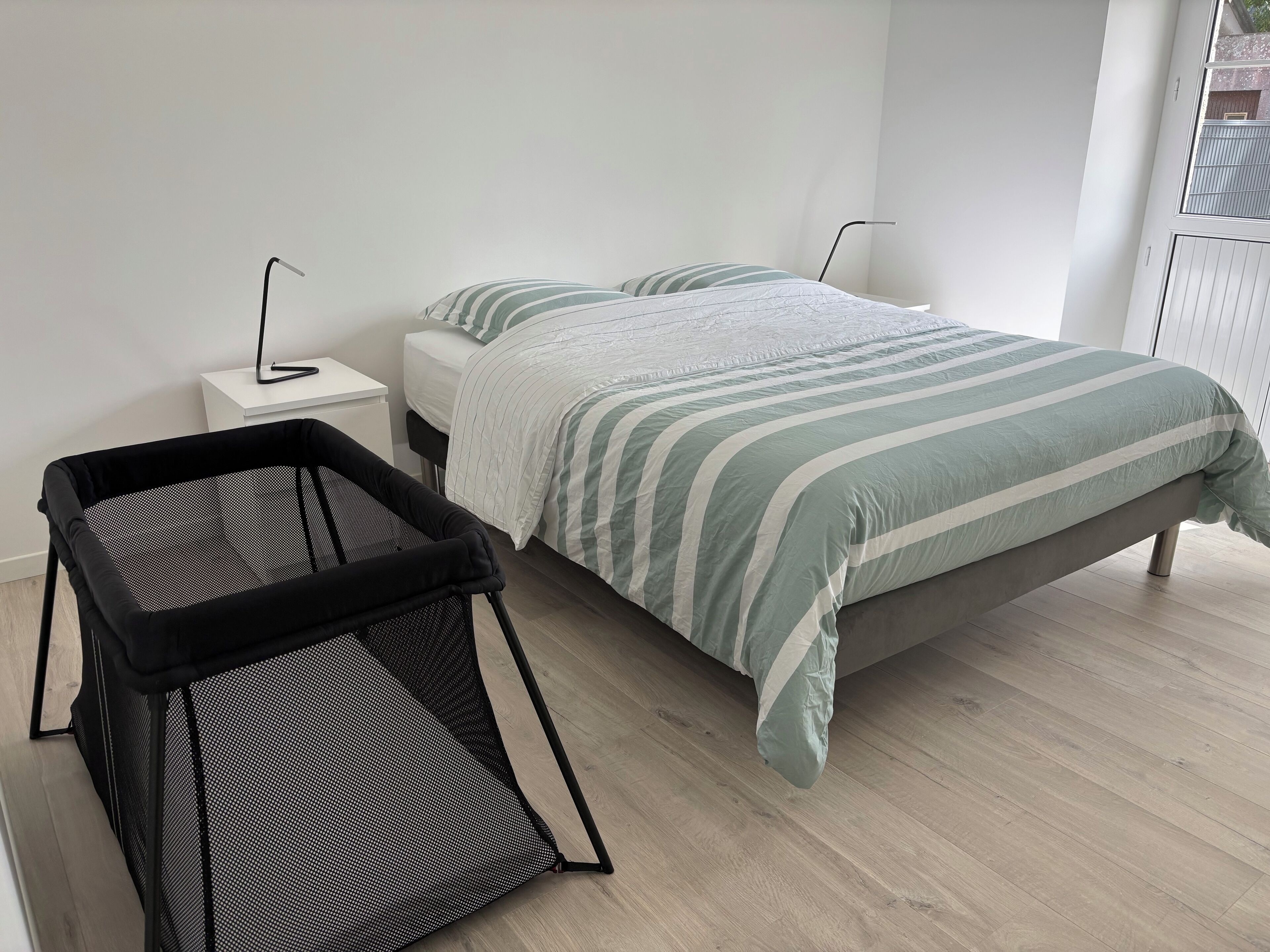 Une chambre à coucher moderne avec un lit, un petit parc pour bébé noir et blanc et une table de chevet blanche avec une lampe.