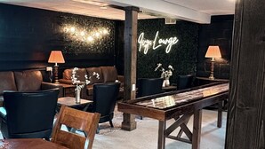 A cozy lounge area with a wooden table, leather seating, and a neon sign reading "Lounge". - Underground Speakeasy Experience in Historic Fort Recovery - Sleeps 14 (Fort Recovery)