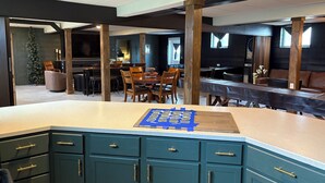 A modern kitchen with teal cabinetry, a central island, and a dining area with wooden chairs and a piano. - Underground Speakeasy Experience in Historic Fort Recovery - Sleeps 14 (Fort Recovery)