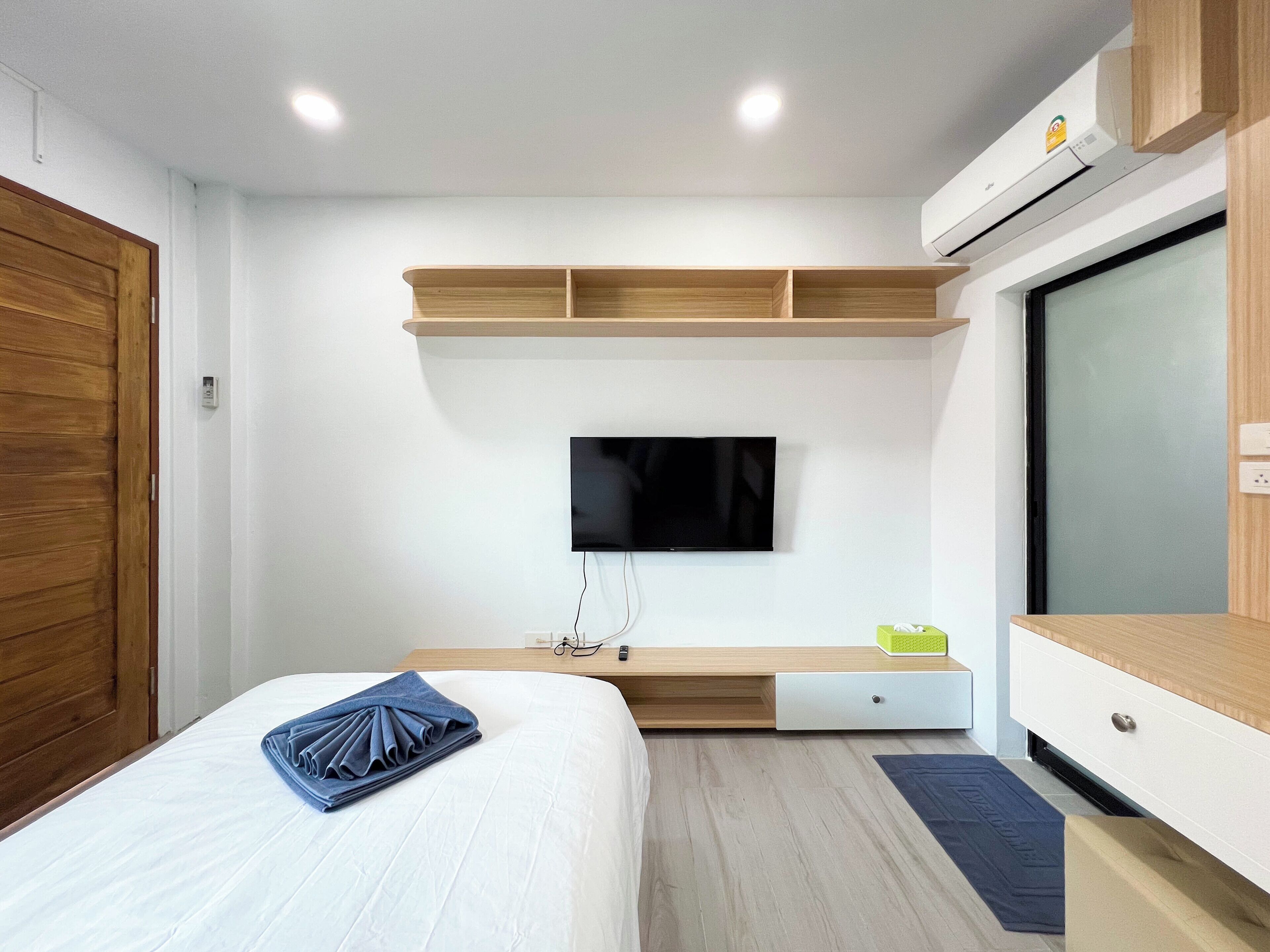 A modern hotel room with a bed, a flat-screen TV mounted on a wooden shelf, a bedside table with a lamp, and a large window.