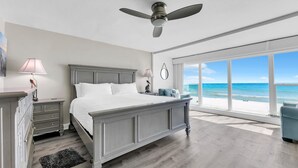 Ein Schlafzimmer mit einem großen Bett, einem Deckenventilator, einem Nachttisch, einer Kommode, einer Lampe und Blick auf den Strand durch große Fenster.