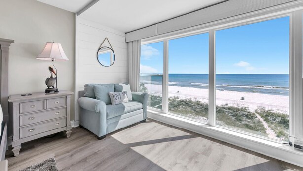 Ein Zimmer mit großem Fenster zum Strand, einem hellblauen Sofa, einer Kommode mit Schubladen und einer Lampe.