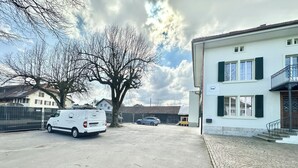 Un área de estacionamiento con una camioneta blanca y un automóvil, un edificio con balcón, y un árbol al fondo.