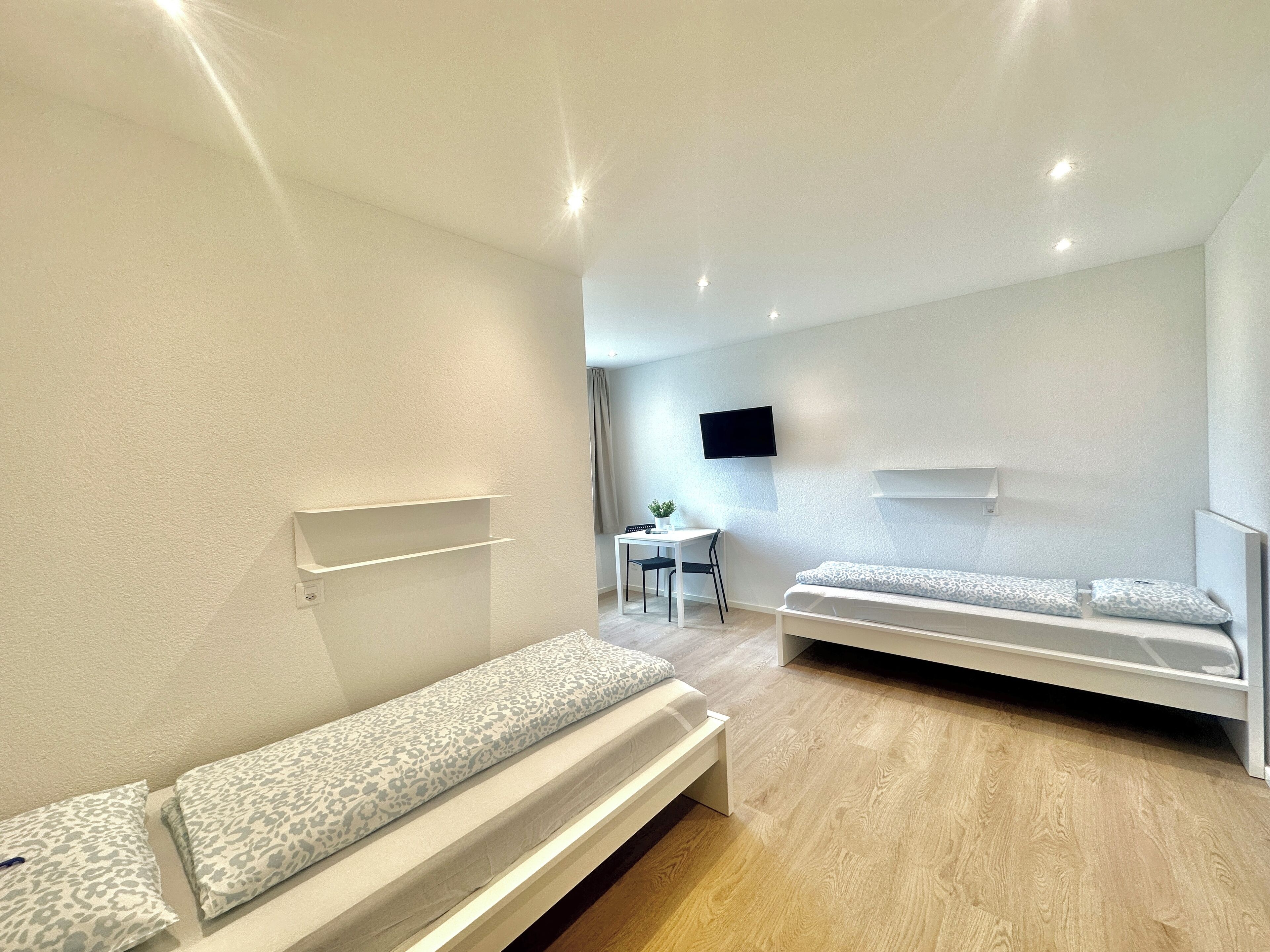 Ein modernes, minimalistisches Zimmer mit zwei Einzelbetten, einem Schreibtisch und einem an der Wand montierten Flachbildfernseher.