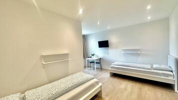 Ein modernes, minimalistisches Zimmer mit zwei Einzelbetten, einem Schreibtisch und einem an der Wand montierten Flachbildfernseher.