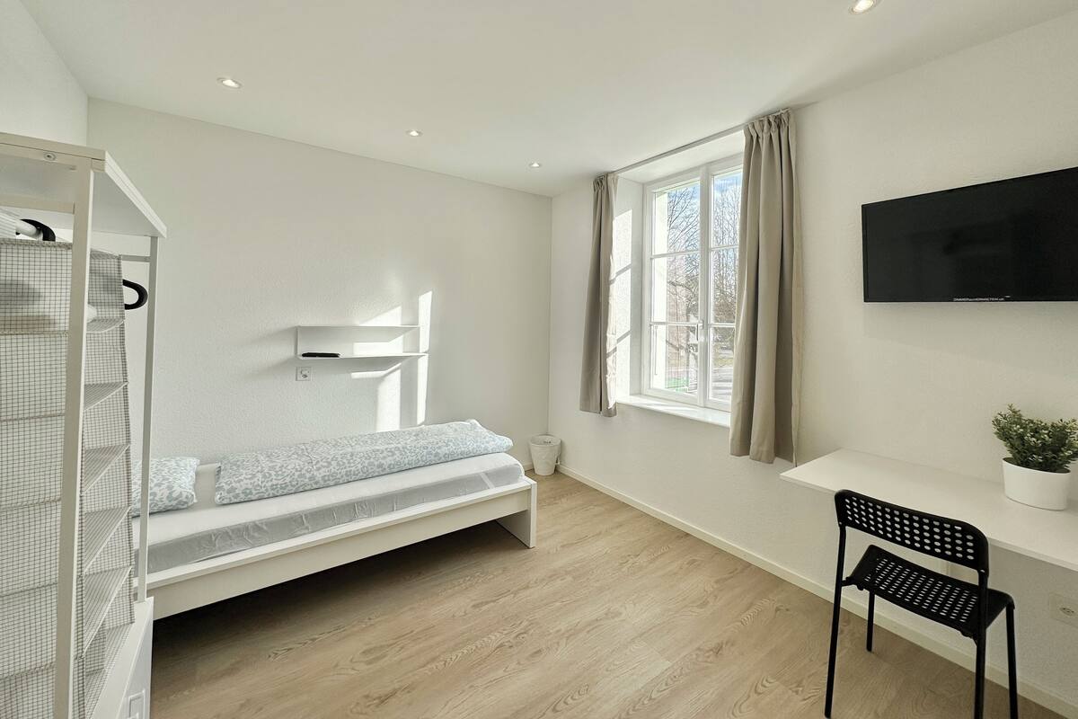 A modern bedroom with a bed, a desk, a chair, a TV, and a window with curtains.