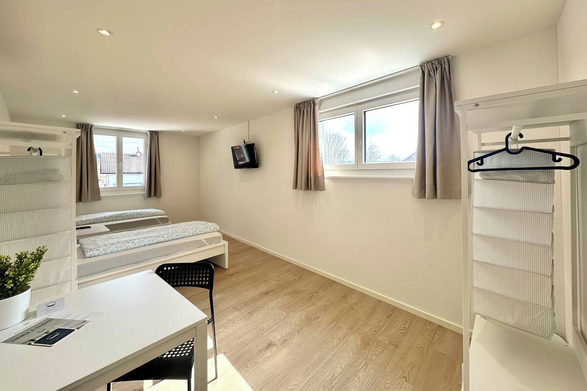 A small, well-lit room with two beds, a desk, a chair, a TV, and a window with curtains.
