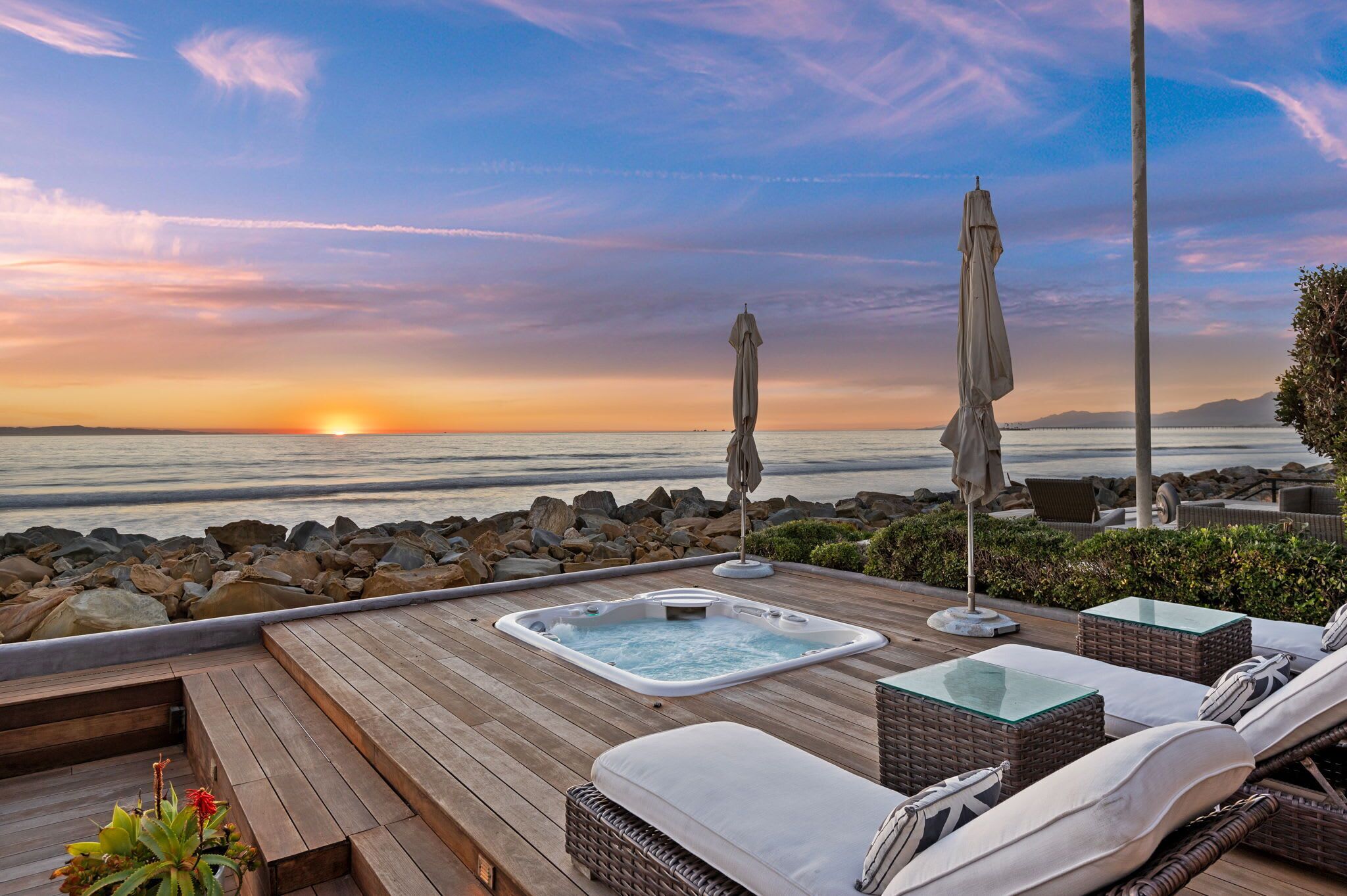 A wooden deck with a hot tub, lounge chairs, and umbrellas overlooking a sunset over the ocean.
