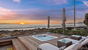 A wooden deck with a hot tub, lounge chairs, and umbrellas overlooking a sunset over the ocean.