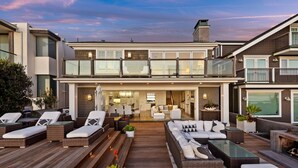 Ein modernes, zweistöckiges Haus mit einer großzügigen Außenpatio-Terrasse, die Korbmöbel, einen Essbereich und eine durch große Fenster sichtbare Küche bietet.