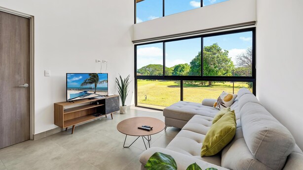 Una sala de estar moderna con un ventanal grande, un televisor de pantalla plana, un sofá, una mesita de centro y un mueble de entretenimiento de madera.