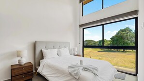 Un dormitorio moderno con un ventanal grande, una cama con sábanas blancas, una mesita de noche con una lámpara y vistas a un paisaje verde.