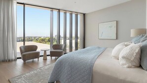 A modern hotel room with a large bed, two armchairs, a small table, and a view of the city through floor-to-ceiling windows. - Luxury Villas Como Penthouse (Melbourne skyline and river views) (South Yarra)