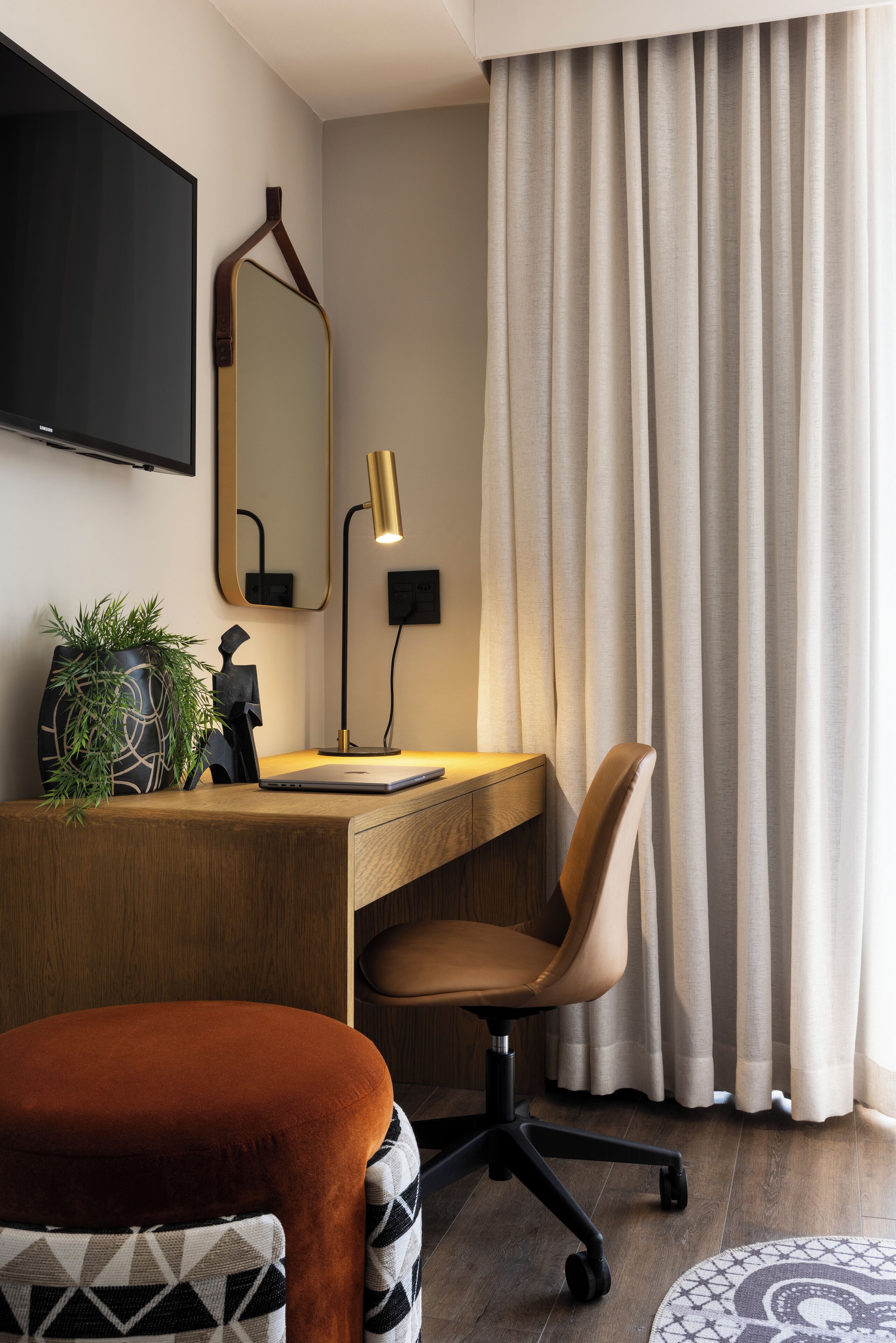 Moderni hotellihuone, jossa on puinen työpöytä, kannettava tietokone, tuoli, ruukussa oleva kasvi, peili ja suuri ikkuna, jossa on verhot.