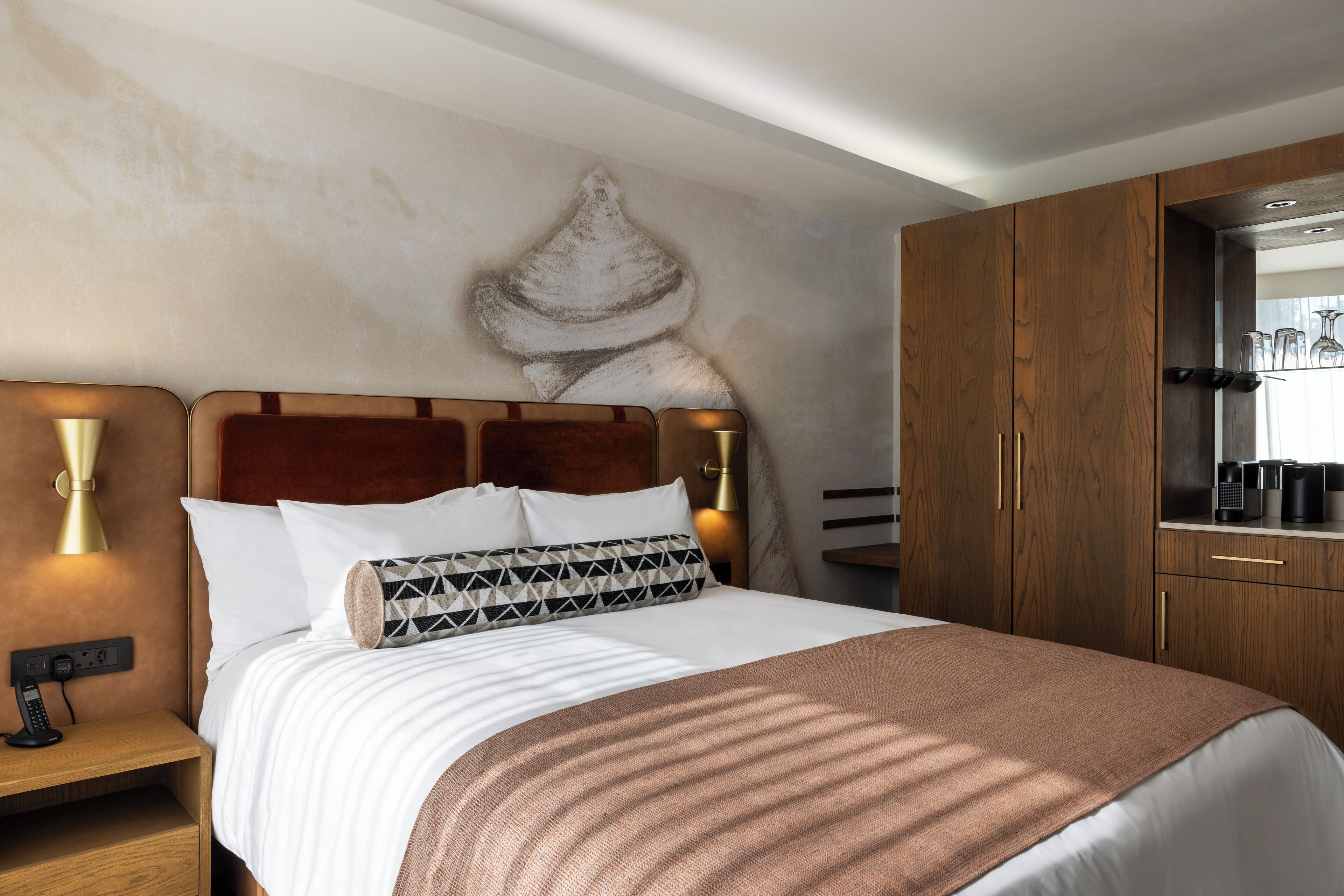 Habitación de hotel con cama, cabecero de madera, lámparas de noche, mesita de noche y minibar.