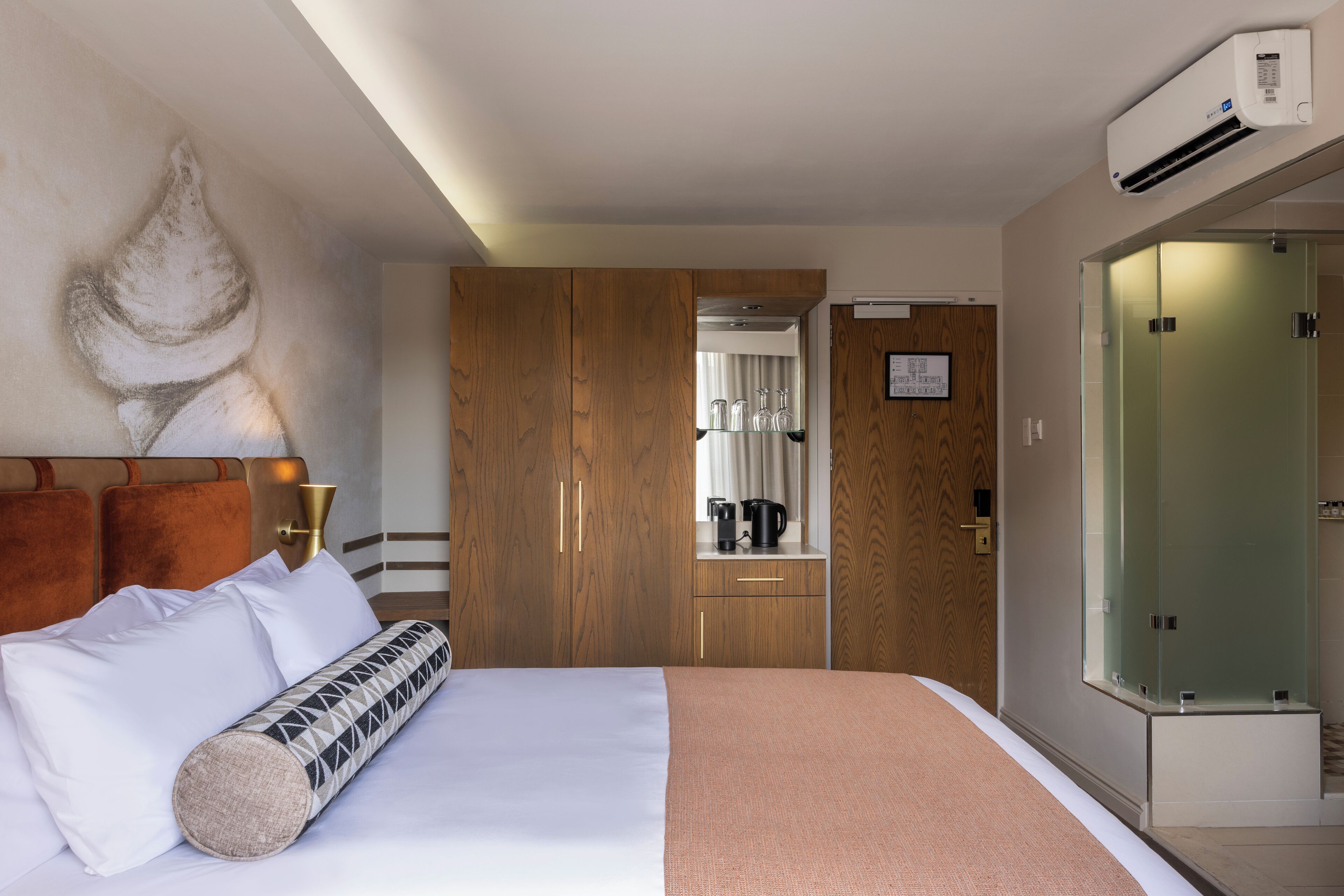 A hotel room with a bed, a wooden wardrobe, a glass shower enclosure, and a coffee machine.