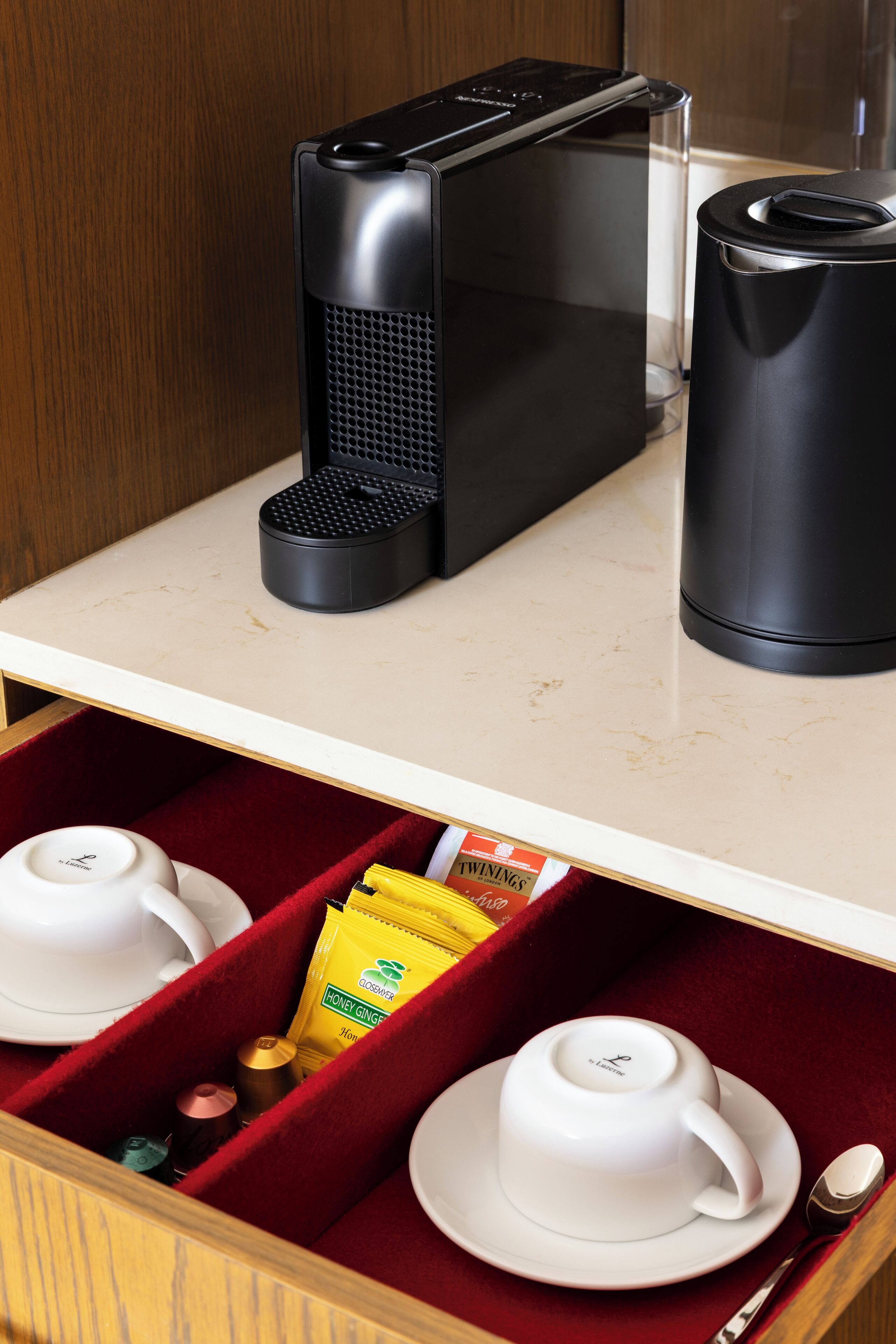 A kitchen counter with a coffee maker, a kettle, and a drawer containing cups, a spoon, and a packet of honey ginger.