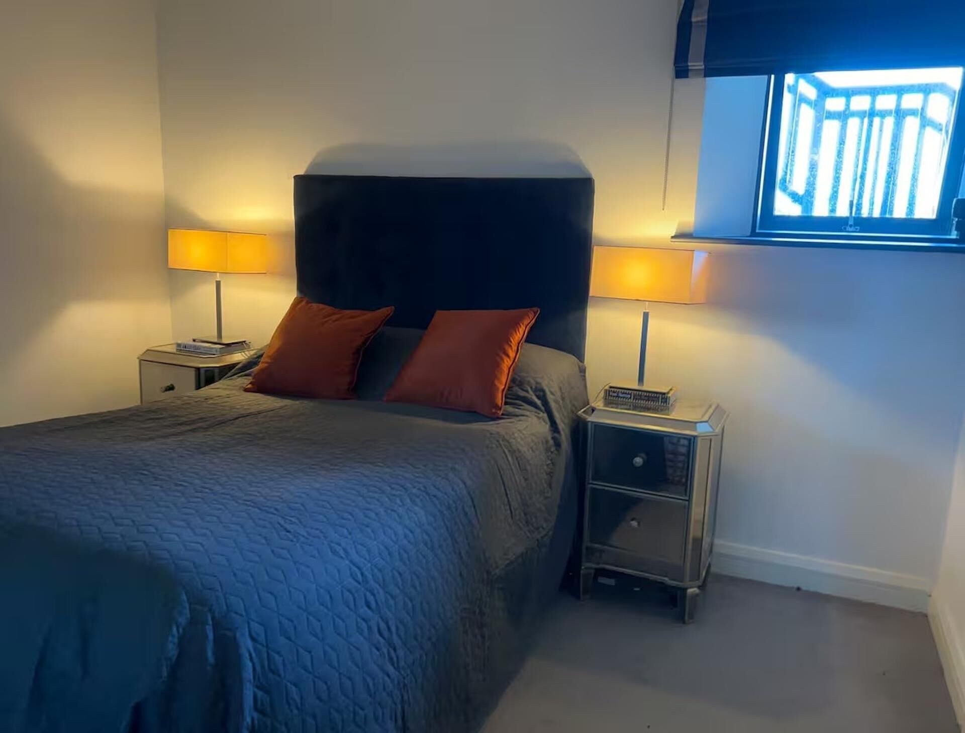 Un dormitorio con cama, dos mesitas de noche, dos lámparas y una ventana.