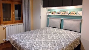 Ein Schlafzimmer mit Bett, einem Fenster mit Vorhängen, einem Heizkörper und einem Regal mit gerahmten Fotos.